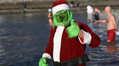 Getty Images A person dressed up as the Grinch wearing a father Christmas suit, they put their thumb up at the camera. They are submerged to their waist in the sea.