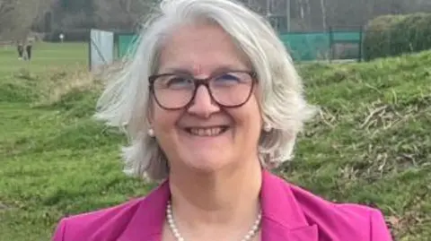 Cambridgeshire Fire and Rescue Head and shoulders photo of Edna Murphy in a park with grass fields around her. She wears a pink blazer, white pearl necklace, matching earrings and glasses. She has grey hair which reaches just above her shoulders and is smiling at the camera. 