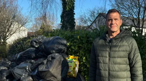 A man with dark hair and a green coat next to a pile of black bin bags. He looks into the camera while next to him are several bin bags which come to the top of a hedge behind the bags and himself. Behind the hedge are several trees.