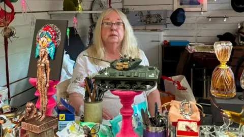 LDRS A woman with long blonde hair, glasses and a white short sleeved linen top looks concerned as she sits behind her market stall. A range of ornaments including an Egyptian woman and a model of an army tank, as well as some items of jewellery can be seen in front of her, with a picture of a zebra behind her.