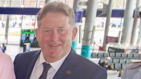 PA Media Darragh is wearing a navy suit with a pin on the lapel. He is around 60 and his hair is greyish and parted in the middle. 