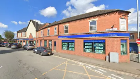 A brick building by the side of a road with several cars parked next to it. The blue branding of an estate agent is on the ground floor.