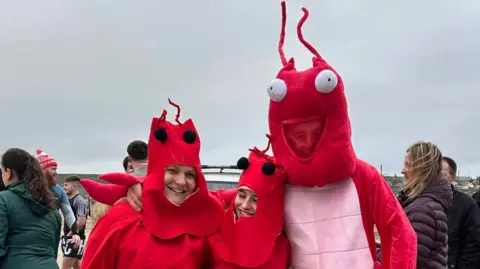 Peter Sawyer Three people wearing lobster fancy dress costumes standing on the beach with people walking around in the background.