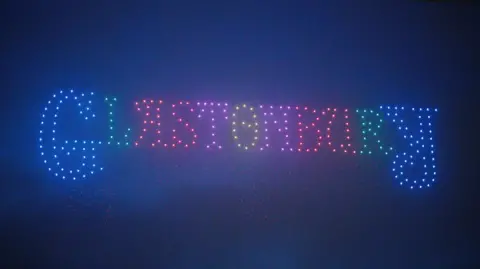 PA Media Image of a drone show at Glastonbury Festival. Glastonbury is spelled out with different coloured lights.