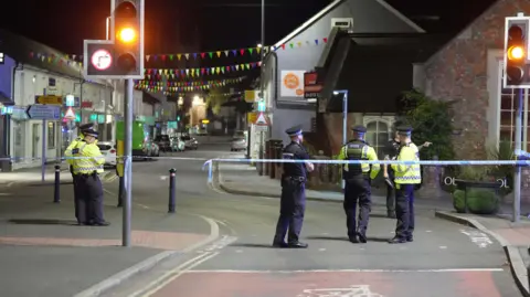 Eddie Mitchell Police officers stand on the road and pavement behind a cordon at night near a town high street. There is bunting strung between the buildings .