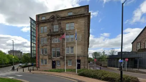 A five-storey building with a glass-panelled side with flags flying outside