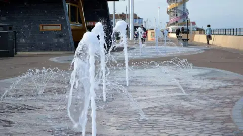 Multiple jets of water shooting up out of the ground on the beachfront location. There is a red/cream mosaic design under the water streams. There are railings overlooking the sea on the right and a tower with a swirling pink and yellow design on the exterior in the background.