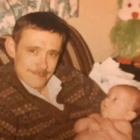 Coutts family Ean Coutts when he was younger sitting on a seat while holding a baby. He has short hair and a moustache and is wearing a patterned brown, v-necked jumper with a shirt underneath.