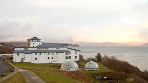 The college is a complex of white-walled buildings with grey roofs. The tallest is a turret like building. Next to the buildings are two small greenhouse-like glass domes. The site is on the coast and overlooks the sea.