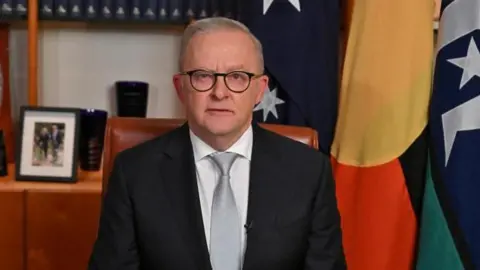 Prime Minister Anthony Albanese delivering a pre-recorded address to the nation in his office at Parliament House, Canberra. He wears a dark grey suit and pale grey tie. The office he sits in is wooden and behind him there are several flags hanging and picture frames.