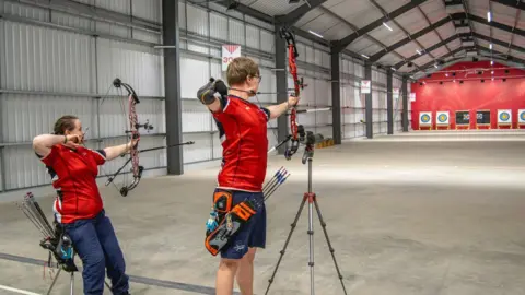 Archery GB Two archers in red tops and blue shorts aiming at targets at the far end of a long sports hall.