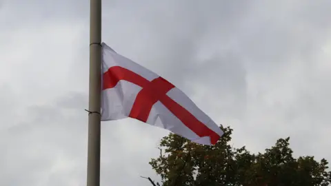 BBC A St George's flag attached to a lamp-post with a grey sky and the top of a tree in the background.