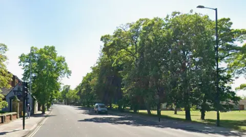 Marton Road has four narrow lanes. It is surrounded by houses on its left side and grassland on the right. The road is lined by trees. A small grey van is travelling down towards a set of traffic lights.