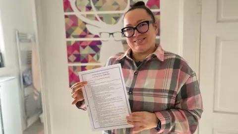 Agnieszka Barys wearing a pink and grey checked shirt. She is standing in front of a white wall which has a canvas on it with a white deer wearing black glasses. Agnieszka has dark hair pulled back into a bun and is wearing dark-rimmed glasses. She is smiling. She is holding a white piece of paper.