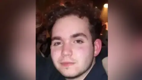 A head and shoulders photograph of Connor. He has dark wavy hair and is standing in a dark room with a spotlight in the background.