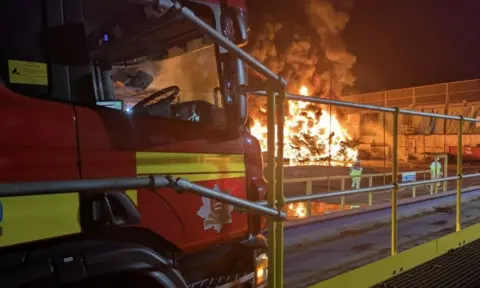 Essex Fire and Rescue A scene at night. The front of a red fire engine in the foreground, with a large fire and smoke in the background. It is almost white with heat. Two fire fighters seen from behind with helmets and protective gear on. There is a big industrial building or warehouse next to the fire, which is in the open.
