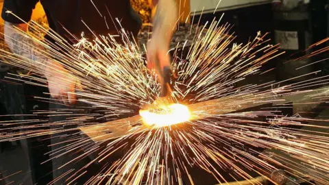 A Blacksmith smashes a horse shoe, which is glowing yellow with heat, with a hammer causing sparks to fly off it. The craftsperson is wearing a black shirt and has tattooed arms.