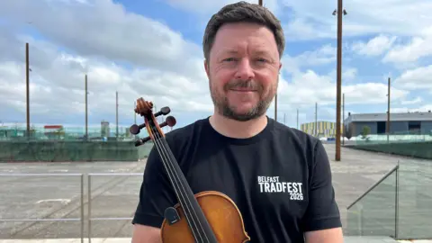 Dónal is holding a fiddle and smiling. He's wearing a black top that says Belfast TradFest 2026. The Titanic slipways are in the background, with metal poles lining the space. It's a cloudy day. 