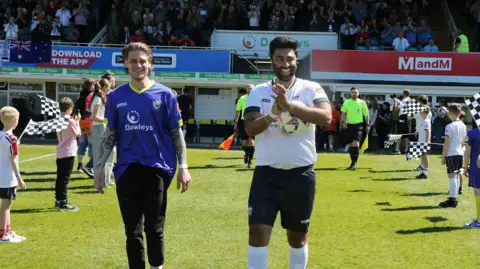 Steve Niblett Rai wearing a full Hereford FC kit walks onto the Edgar Street pitch holding the match ball alongside another man wearing a purple Hereford FC top and black trousers. 