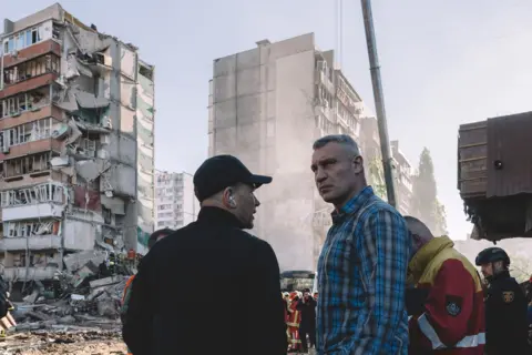 Andrew Kravchenko / Bloomberg via Getty Images Two people stand in front of rescuers with damaged residential buildings and rubble in the background on a sunny day in Kyiv on Thursday.