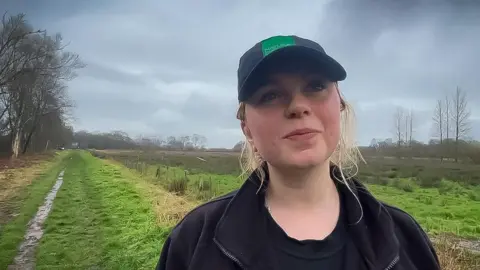 A young woman in a Natural England cap standing in front of a grassy lane that leads into the nature reserve