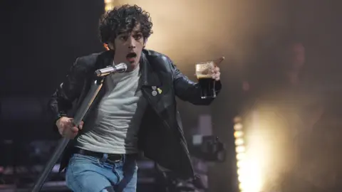 Matty Healey is on stage wearing a black leather jacket. He is walking towards the camera holding a microphone and a glass in each hand. He has black, curly hair.
