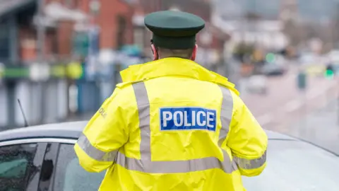 A PSNI police officer wearing a high-visibility jacket closes off a street. Blue and white police tape is up. The street in the background is out of focus.
