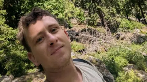 Submitted A man with short brown hair which is slightly curly on top taking a selfie surrounded by rocks, trees and general greenery. He is wearing a beige top and a black backpack.