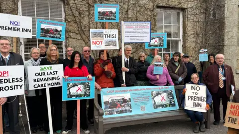 Lee Trewhela/LDRS About 20 people outside a hotel holding placards and banners against the proposed plans. There is a printed banner attached to a bench with a lady sat next to it with a banner saying 'No to Premier Inn'.