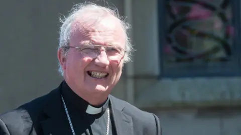 Pacemaker a smiling Bishop Donal mckeown in full clerical dress standing in front of a stained glass window