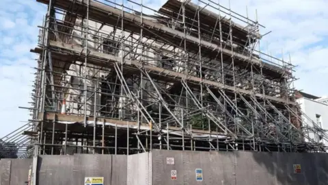 A grey building covered in a large quantity of scaffolding and surrounded by grey hoardings