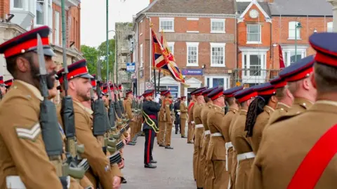 Maj Chris Brown A view of personnel holding flags in between rows of soldiers in Khaki uniform holding weapons.