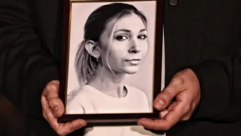 Imagens globais Ucrânia via Getty Images Uma mulher tem uma foto em preto e branco de Viktoriia Roshchyna, que a mostra olhando direto para o barril da câmera, e seus cabelos cansados em um rabo de cavalo.