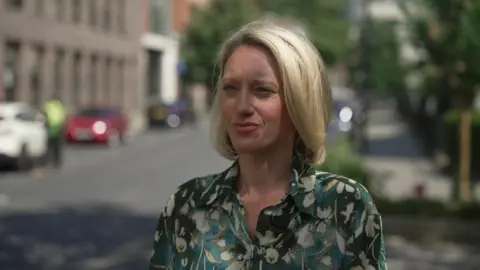 Claire Waxman, with shoulder-length blonde hair and wearing a dark green shirt with white flowers on it is standing on a London street