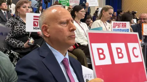 BBC correspondent Steve Rosenberg is seen at a press conference holding a large red sign saying "BBC"