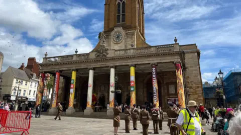 A large brick church with eight stone columns connection the stairs to a balcony. A parade marches in front of the church. 