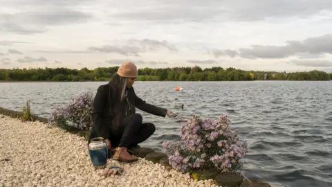 Getty Images Woman at side of water on pebble stone path and by water and with urn by her side scattering ashes