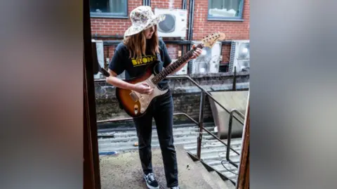 Sonja Sleator is playing an electric guitar. It's brown and white. She's wearing a hat with a flower pattern, a top that says the Bronze on it and jeans and trainers. A brick building is behind her. She's standing outside of a doorway. 