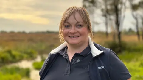 Samantha McCaroll has dark blond hair tied back and is smiling. She is wearing a polo-top with a body warmer over it and is standing in front of peatland