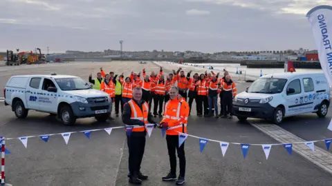 Two people in orange fluorescent jackets cut a ribbon decorated with blue and white flags, with two vans and group of cheering people in the background