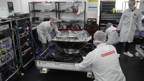 Three engineers wearing lab coats, head coverings and mask work around a satellite on a trolley.