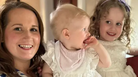 Kim Gawley A woman with dark brown hair is looking at the camera and smiling. She is holding a blond baby in her arms, the child is wearing a cream top, pink bib and has her hand in her mouth. Beside her is another young child with shoulder-length dark blonde curls. She is wearing a cream dress and looking off to the side of the image. 