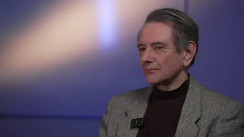 Matt Wingett has short brown hair, and is wearing a grey blazer over a burgundy turtleneck.