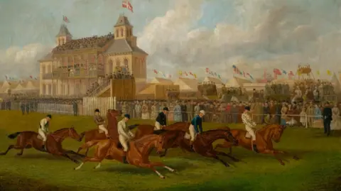 A Victorian painting showing six horse riders racing past a grandstand and large crowds