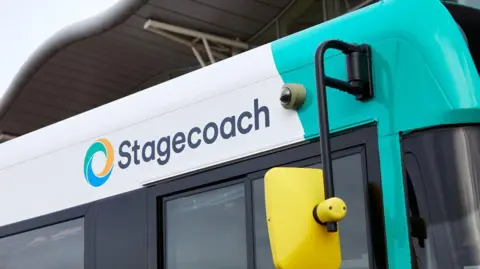 The side of a Stagecoach bus with its logo next to a multicoloured circle with a very bright yellow wing mirror in the foreground