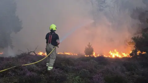 Suffolk Fire and Rescue Service Firefighter sprays liquid at fire through yellow hose. The fire burns heathland in front of them, with smoke rising in to the air