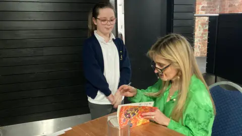 A woman with long blonde hair and black-rimmed glasses wearing a bright green shirt is sitting on a blue chair and signing a copy of her book for a young school pupil, who is wearing a white T-shirt and blue school cardigan.