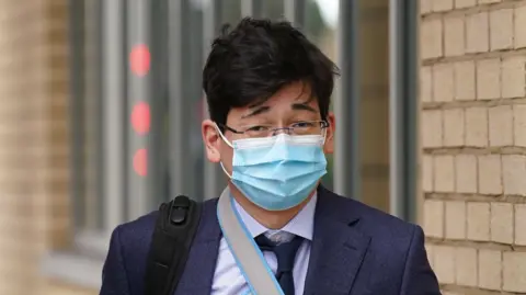 Jonathon Dean wearing a navy suit with a navy tie and a white shirt and glasses. He is walking outside of a court building with the strap of a black rucksack over one shoulder and a facemask on.