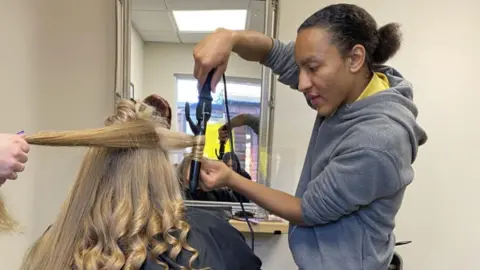 Cameron with dark hair tied at the back wearing a grey top winds hair around some straighteners in front of a mirror. The long blonde hair, with curls at the bottom, belongs to a woman with her back to the camera.