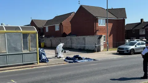 A man dressed in white overalls is collecting items from the crime scene next to a yellow framed bus stop. A policeman is standing in the road which is closed off.
